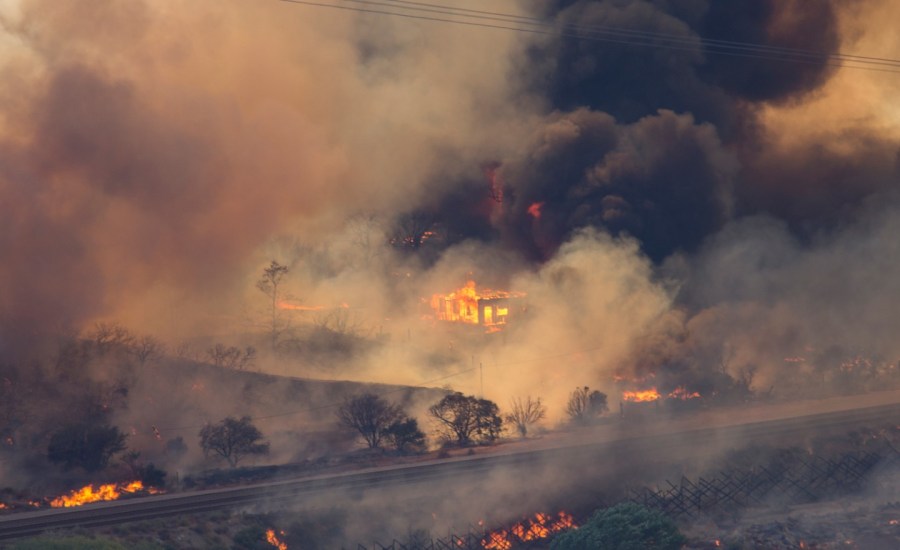 Dark smoke billows all around as a structure and the surrounding landscape are on fire.