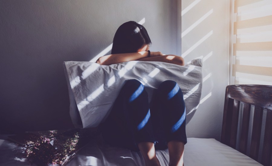 a person sitting on a bed buries their face into a pillow in a dark room with scant daylight coming in through the blinds of an adjacent window