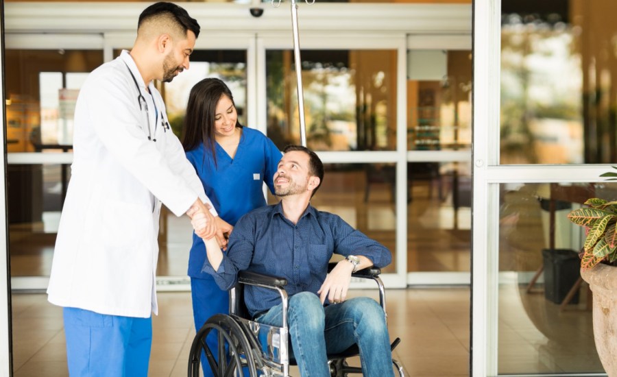 a young female nurse pushes a young male patient out of the hospital on a wheelchair, stopping to speak to a doctor