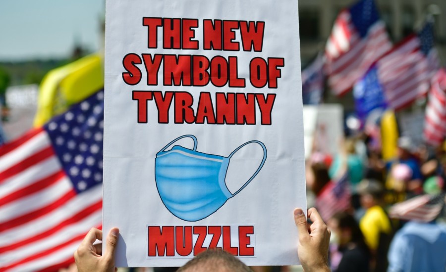 a person at a protest holds a sign that says "the new symbol of tyranny" with a photograph of a medical face mask