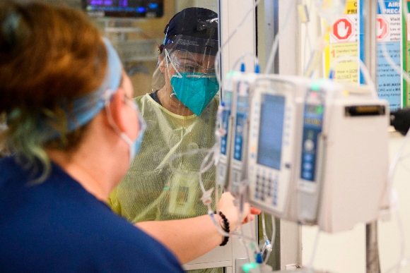 A nurse wearing personal protective equipment (PPE) communicates through a glass door while attending to a patient in a Covid-19 intensive care unit