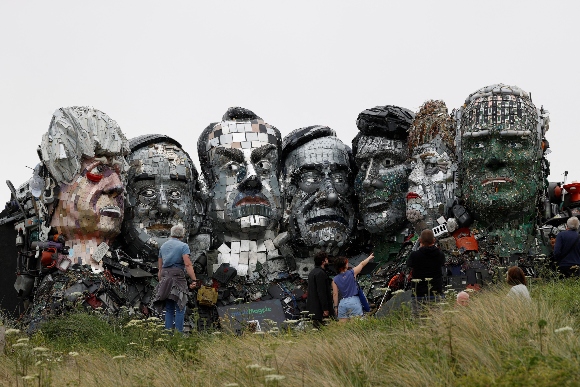 People look at a giant Mount Rushmore-style sculpture of the G7 leaders heads, made entirely of discarded electronics