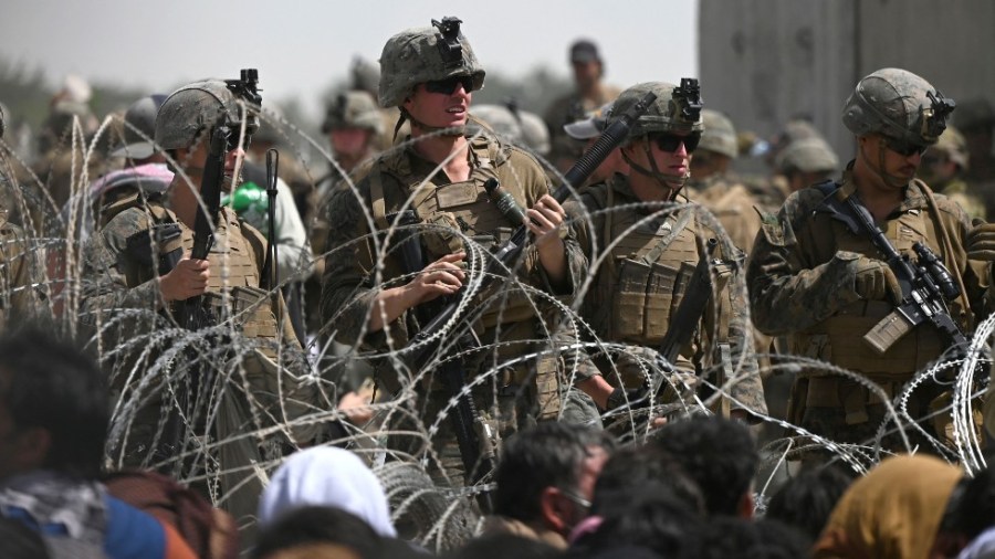 U.S. soldiers stand guard behind barbed wire as Afghans sit on a roadside near the military part of the airport in Kabul