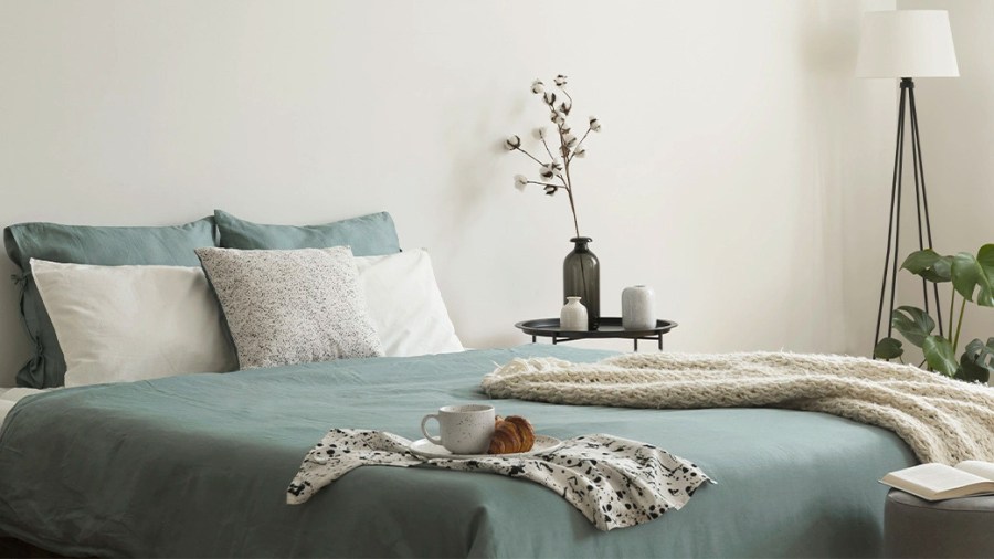 Bed made up with sage green sheets and pillowcases, along with off-white pillows and a throw