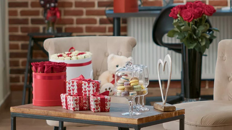 Valentine's Day gift assortment, including white and red roses, a white teddy bear, a heart sculture, macarons, and three gifts wrapped in white paper with red hearts