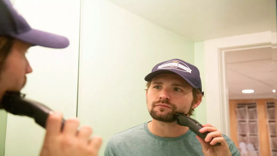 Man wearing a ball cap looking into a mirror while using an electric beard trimmer