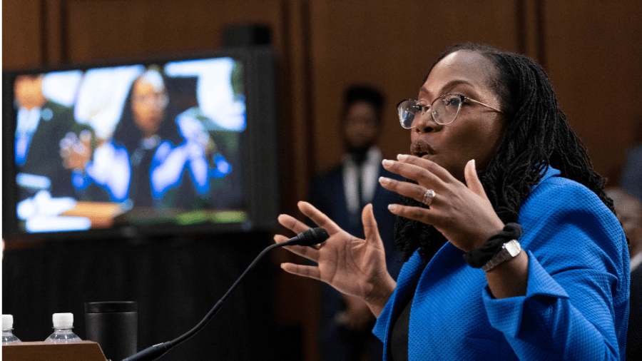 Ketanji Brown Jackson, wearing a blue blazer, holds hands up while speaking. She is seated beside a television screen showing her image.