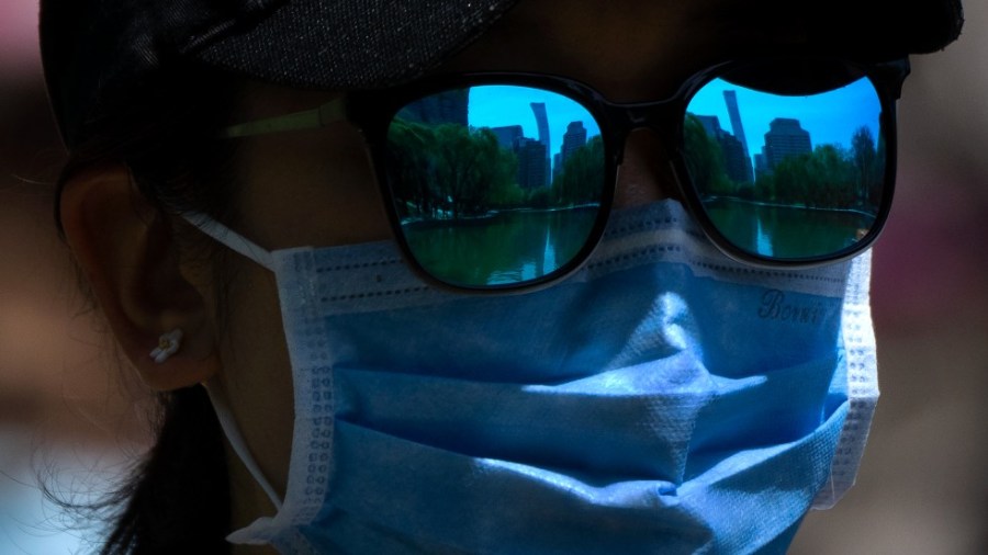 The skyline of Beijing is reflecting in the sunglasses of a woman wearing a face mask as she walks at a public park in Beijing