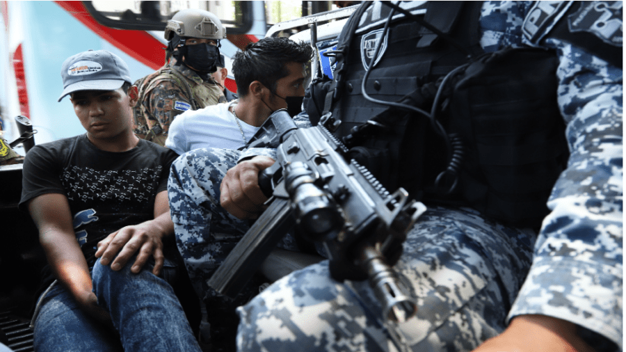 A soldier in blue fatigues, sitting beside detainees, holds a weapon on his lap.