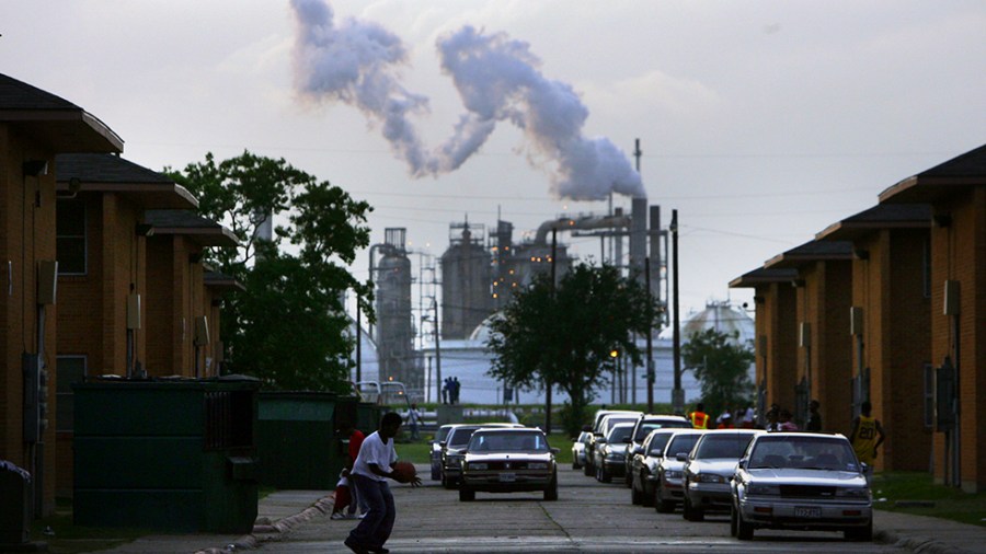 Kids play with a basketball in a street with an oil refinery billowing smoke not far away in the background.