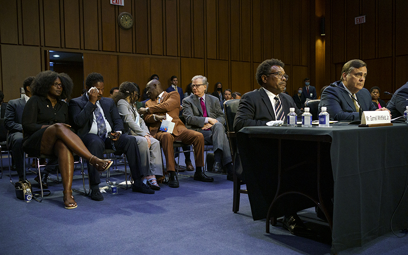 Garnell Whitfield Jr. testifies as hearing attendees are seen conversing in the background