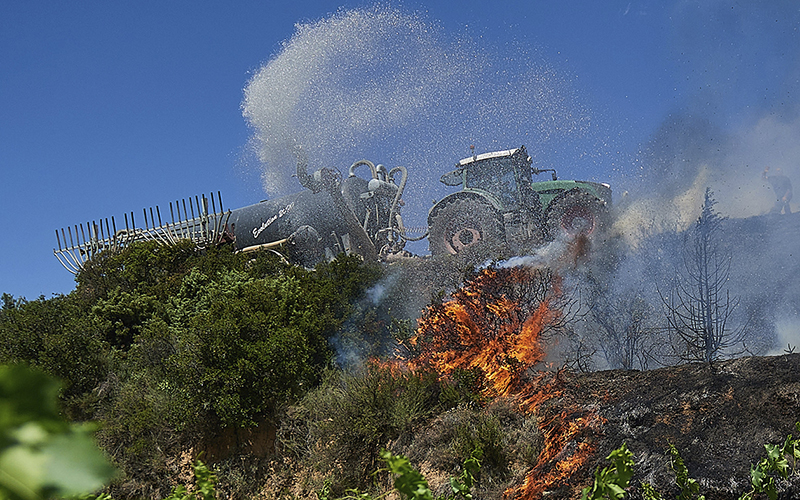 A neighbor with a tractor sprays water on a fire, causing a cloud of mist and smoke to form