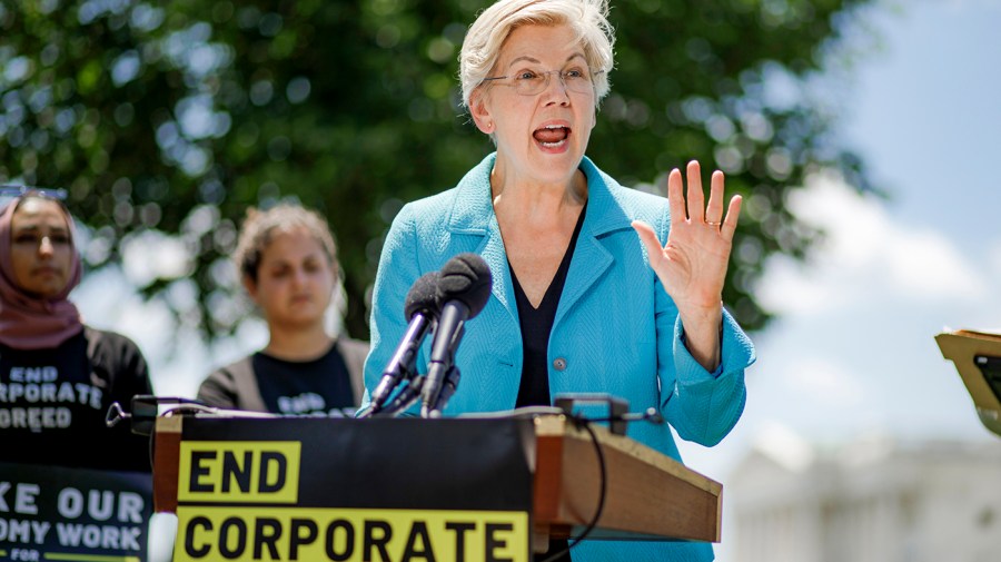 A white woman speaks behind a lectern with a sign that says End Corporate Greed.