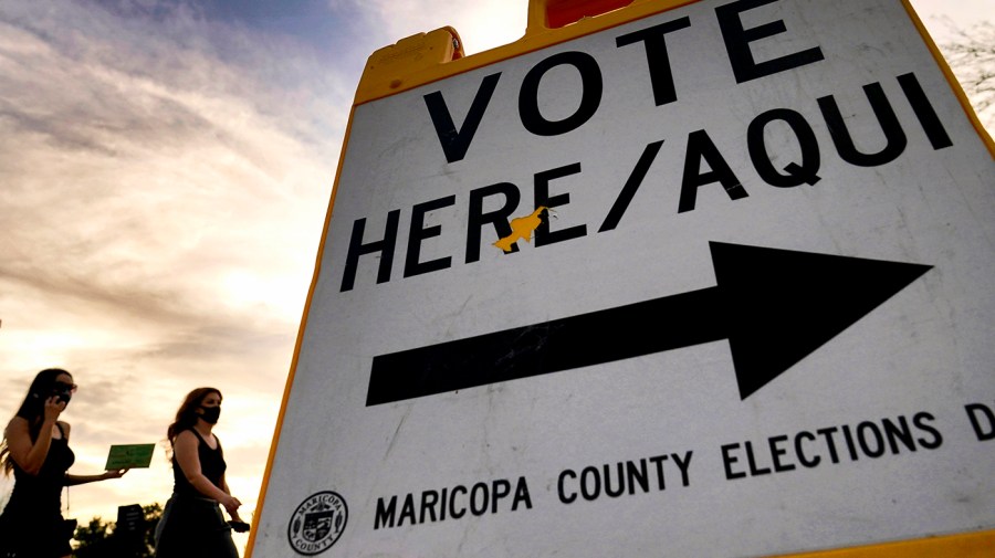 People walk behind a sign that says Vote Here/Aqui.