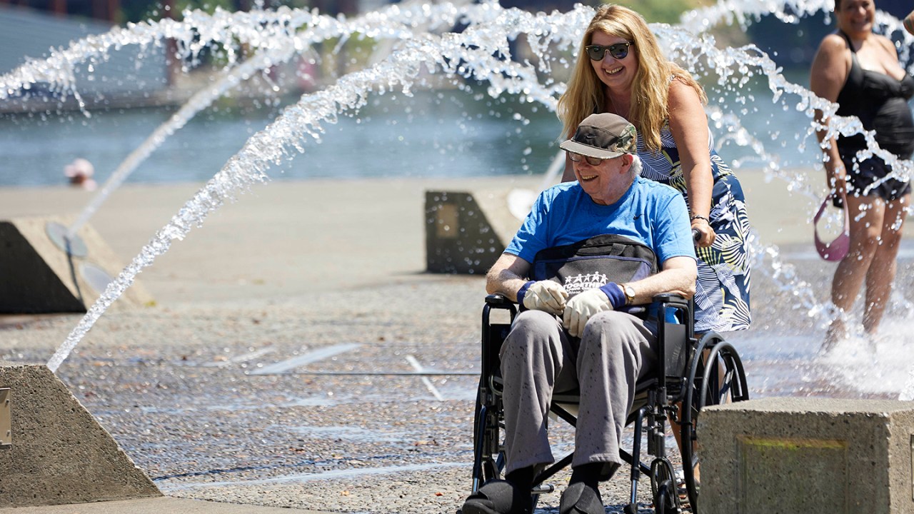 Women pushing man in wheelchair through fountain.