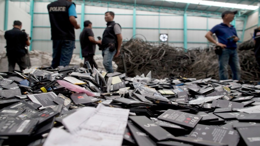 Thai law-enforcement officers and journalist stand behind a pile of mobile batteries during a raid at a factory accused of importing and processing electronic waste, suburbs of Bangkok, Thailand on Thursday, June 21, 2018.