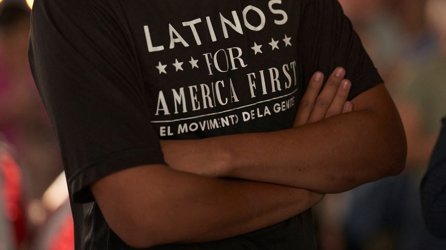 Close-up photo of a person wearing a "Latinos for America First" shirt in white font. Shirt is black