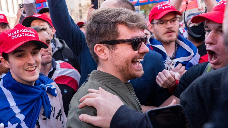 Nick Fuentes smiles while greeting people in a crowd wearing red MAGA hats.