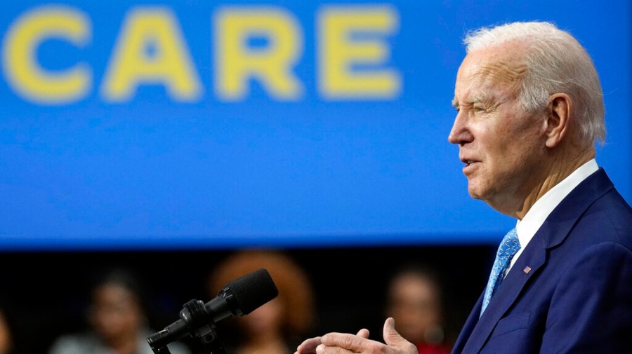 President Biden speaks into a microphone with a bright blue banner in background.