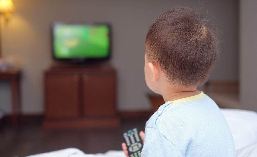 young child sitting on bed facing away at a TV holding a remote controller