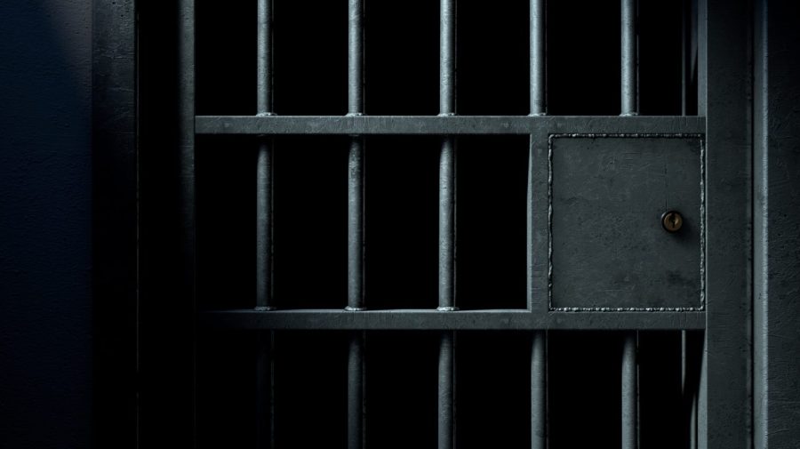 A dark jail cell is seen through a metal bar door.