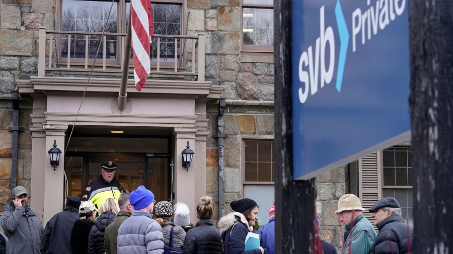 File - A law enforcement official, behind, stands in an entryway to a branch of Silicon Valley Bank, Monday, March 13, 2023, as customers and bystanders line up outside the branch, in Wellesley, Mass.