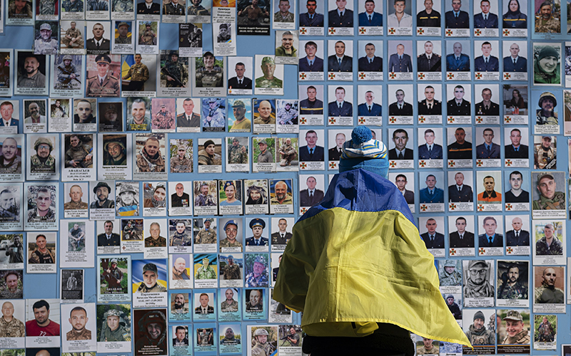 A woman wrapped in a Ukrainian flag stands next to the Memory Wall of Fallen Defenders of Ukraine