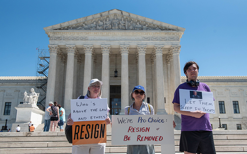 Three protesters hold signs calling out Justice Thomas, with the Supreme Court in the background