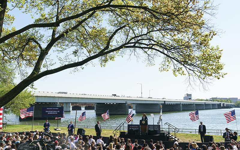 Vice President Harris speaks about bridge projects to a crowd, with branches from a large tree extending overhead and a bridge and river in the background