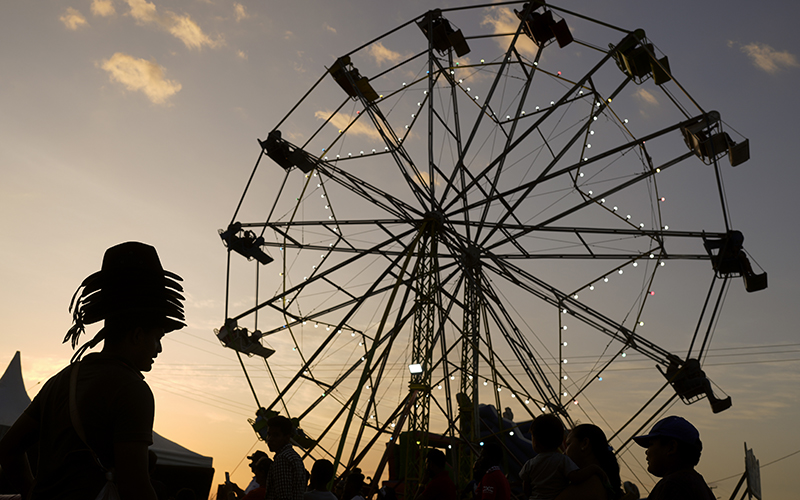A man wearing a stack of hats to sell on his head walks past a ferris wheel during the annual Rupununi Ranchers Rodeo festival