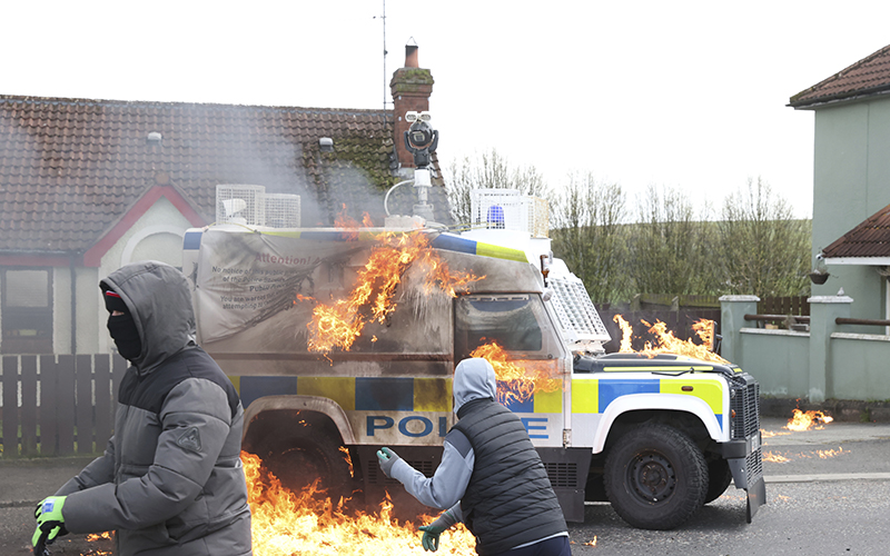 Masked youths throw petrol bombs at a police Land Rover, which is engulfed in flames