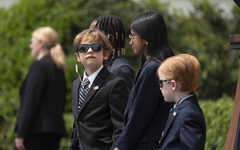 A kid acting as a Secret Service agent, wearing sunglasses, a suit and an earpiece, looks up