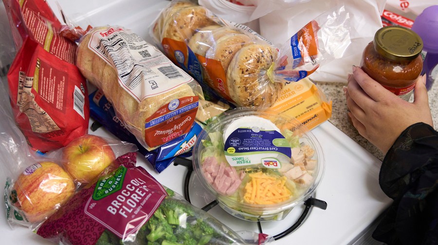 A variety of food items including bagels, bread, pre-made salad and broccoli florets are seen from above as a hand moves on the right-hand side of the photo