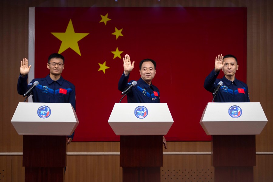 Chinese astronauts for the upcoming Shenzhou-16 mission, from left, Gui Haichao, Jing Haipeng, and Zhu Yangzhu wave as they stand behind glass during a meeting with the press at the Jiuquan Satellite Launch Center in northwest China on Monday, May 29, 2023. China's space program plans to land astronauts on the moon before 2030, a top official with the country's space program said Monday. (AP Photo/Mark Schiefelbein)