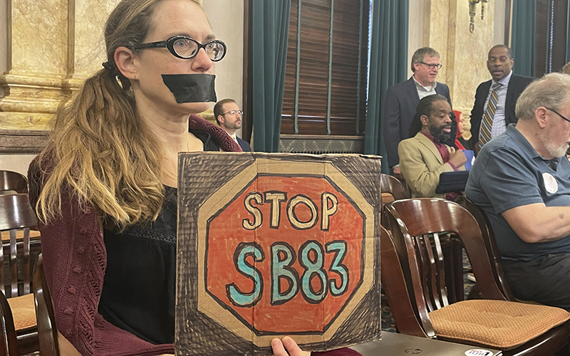 A woman wearing duct tape over her mouth is seen protesting a multifaceted Ohio higher education bill with a sign that reads, "STOP SB83" on a colored red stop sign