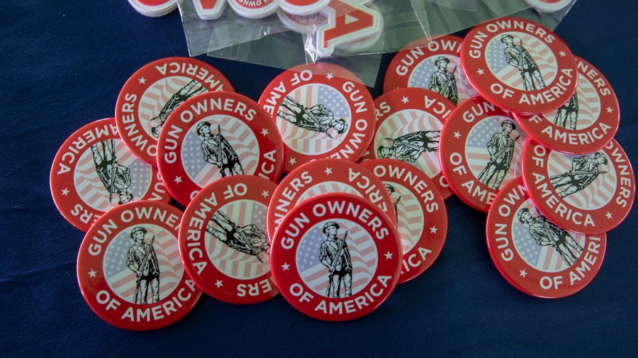 Red and white pins on a blue table read, "Gun owners of America", in caps. In the inner circle of the pin is a colonial man, grayscaled, over a low-opacity American flag.