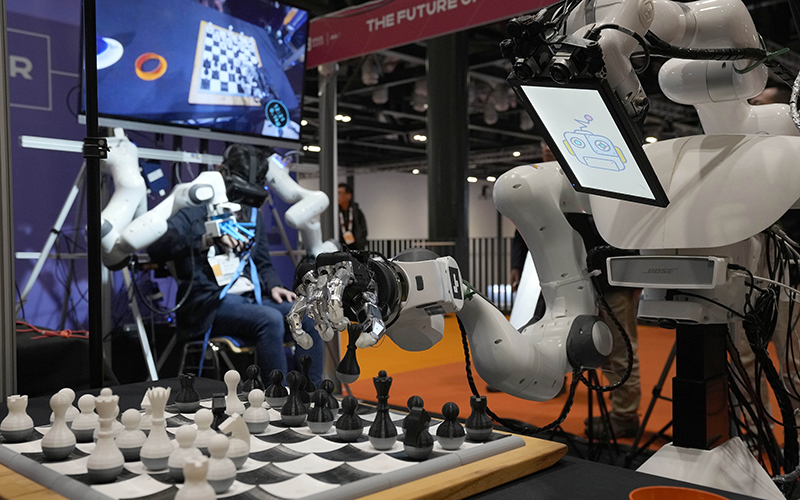 A robot in the foreground makes a chess move while a human in the background controls the robot remotely