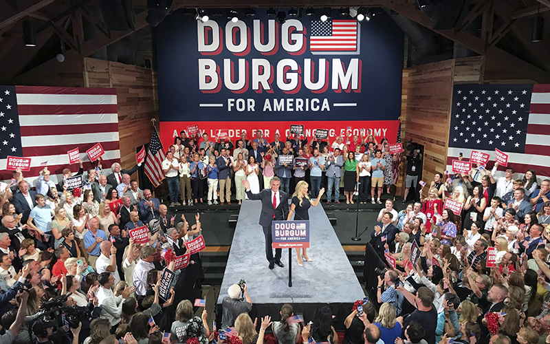 North Dakota Gov. Doug Burgum (R) and first lady Kathryn Burgum stand on a platform in the center of a room full of supporters