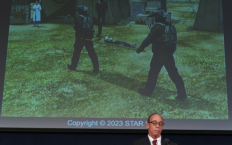 Ufologist Steven Greer stands in front of a projector that shows an illustration of witness testimony from a crash