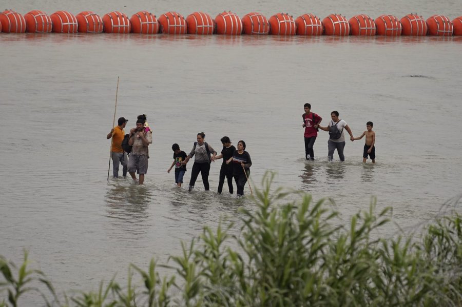 Migrants who crossed the Rio Grande from Mexico walk past large buoys being deployed as a border barrier on the river in Eagle Pass, Texas, Wednesday, July 12, 2023. (AP Photo/Eric Gay)