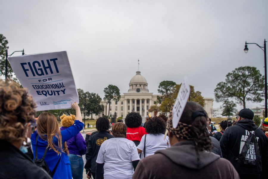 People gather around the Alabama State Capitol holding signs including "Fight for Educational Equity," written in blue letters and in caps on white paper under a cloudy sky