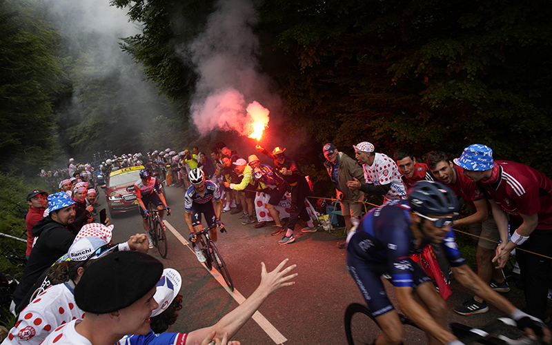 Cyclists in the fifth stage of the Tour de France cycling race along a road lined with supporters, one of whom is holding a lit flare