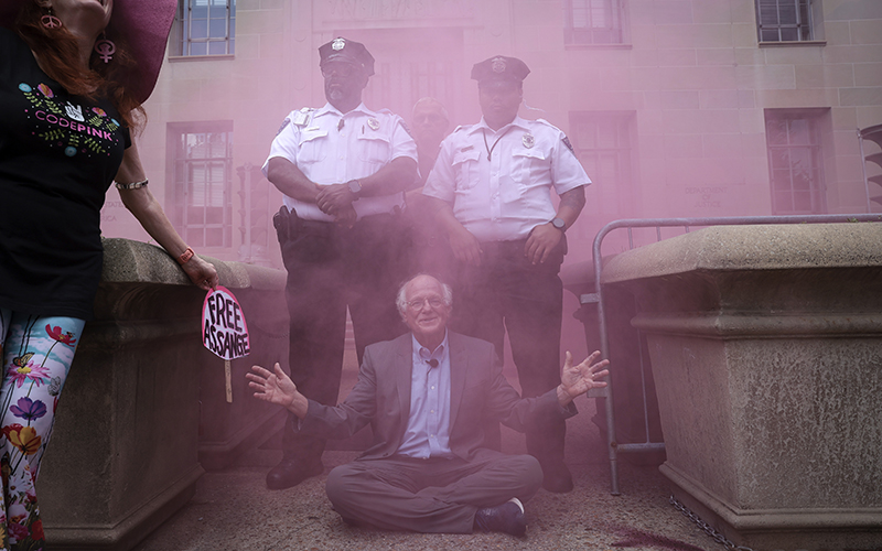 As a pink smoke bomb clouds the air, Ben Cohen, co-founder of Ben & Jerry’s ice cream, sits blocking an entrance outside the Department of Justice as two police officers stand behind him, and another protester holds a sign that reads "Free Assange"