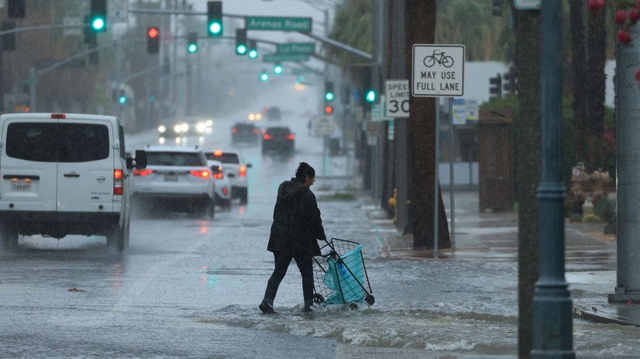 A person pushes a cart on a flooded street as cars drive through water