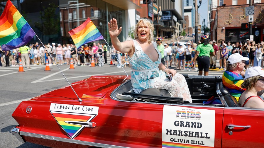 Olivia Hill waves from a red car parade float with rainbow flags and people in the background