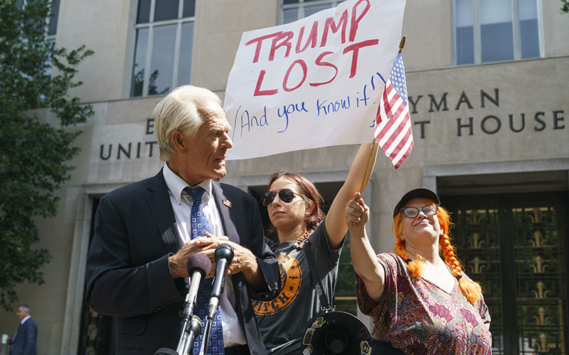 Protesters for and against former President Trump disrupt former Trump advisor Peter Navarro as he speaks to reporters. One of the protesters is holding an American flag, the other is holding a sign that reads "TRUMP LOST (And you know it!)"