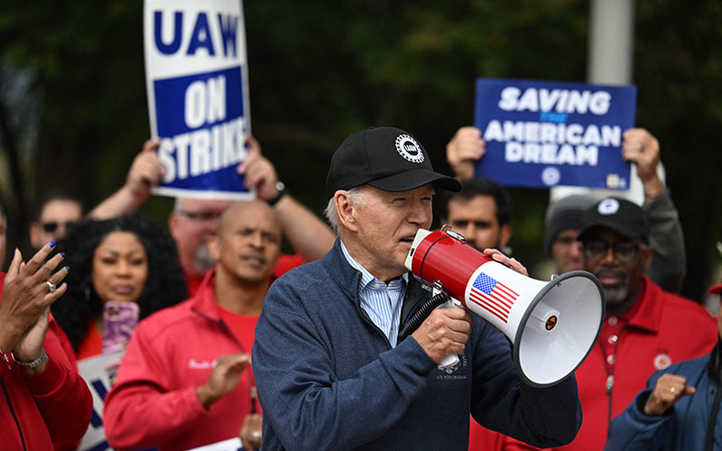 President Biden is in the center of the image, wearing a black UAW baseball cap and speaking into a megaphone that has an American flag on the side. Behind him are members of the UAW, holding signs that read "UAW ON STRIKE" and "SAVING THE AMERICAN DREAM"