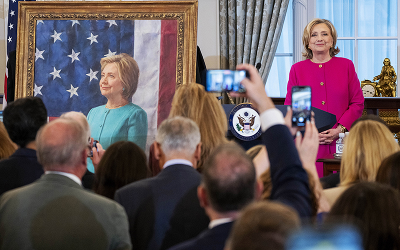 On the right is former Secretary of State Hillary Rodham Clinton, and on the left is her portrait. In the foreground are members of the media holding up their phones to take pictures.