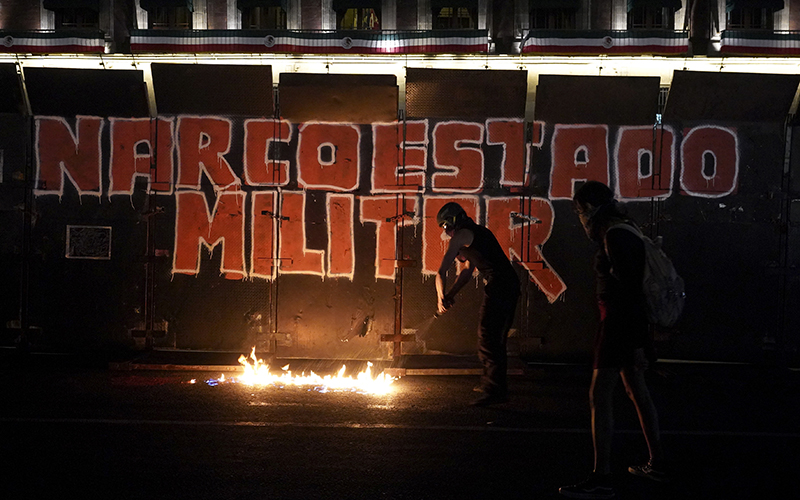 A photo taken at night shows a section of black barriers, on which are the words "ARCOESTADO MILITAR." In front of the barrier, a person in black clothes and a helmet stands by a row of flames.