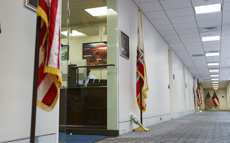 A hallway is seen outside of the doorway to an office. Beside the doorway are two flags: the United States flag on the left side of the door, and the California state flag on the right side. At the base of the flag on the right is a small bouquet of flowers.